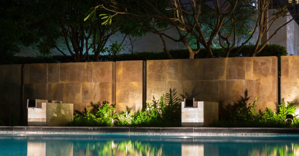 A backyard luxury pool at nighttime with solar spotlights shining on the barrier wall behind the landscaping.