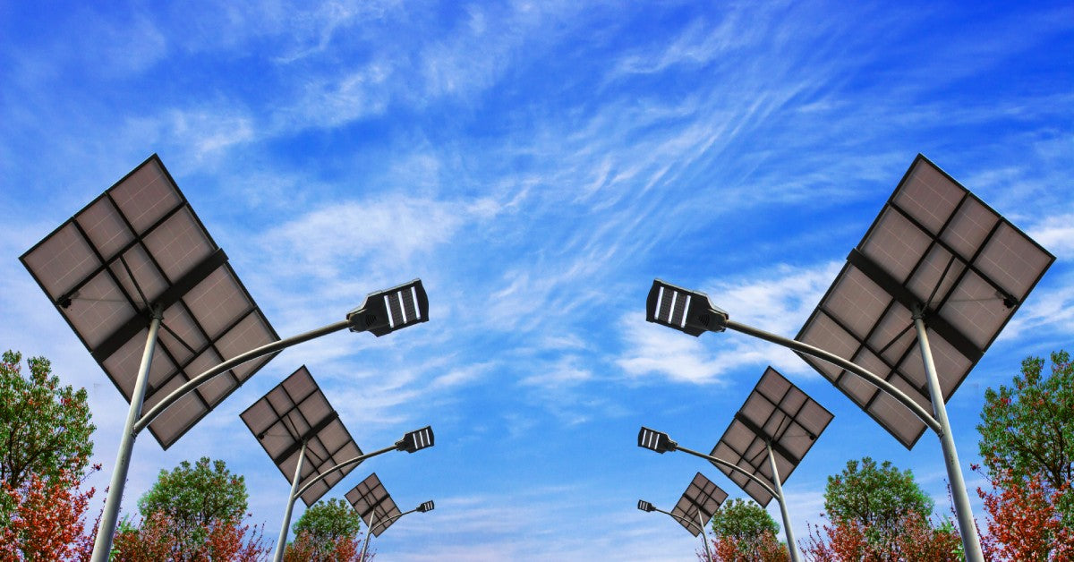 Six solar street lights stand up high against a background of a clear blue sky. Trees with red and green leaves are nearby.