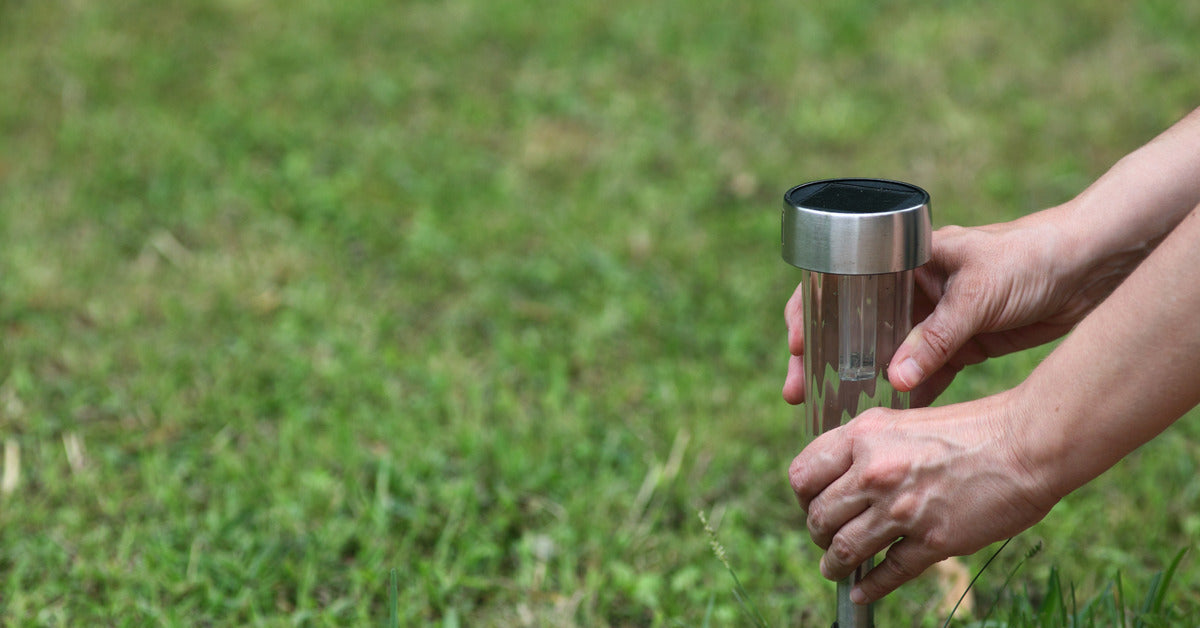 A pair of hands installs a solar pathway light into the ground. A large area of green grass is in the background.