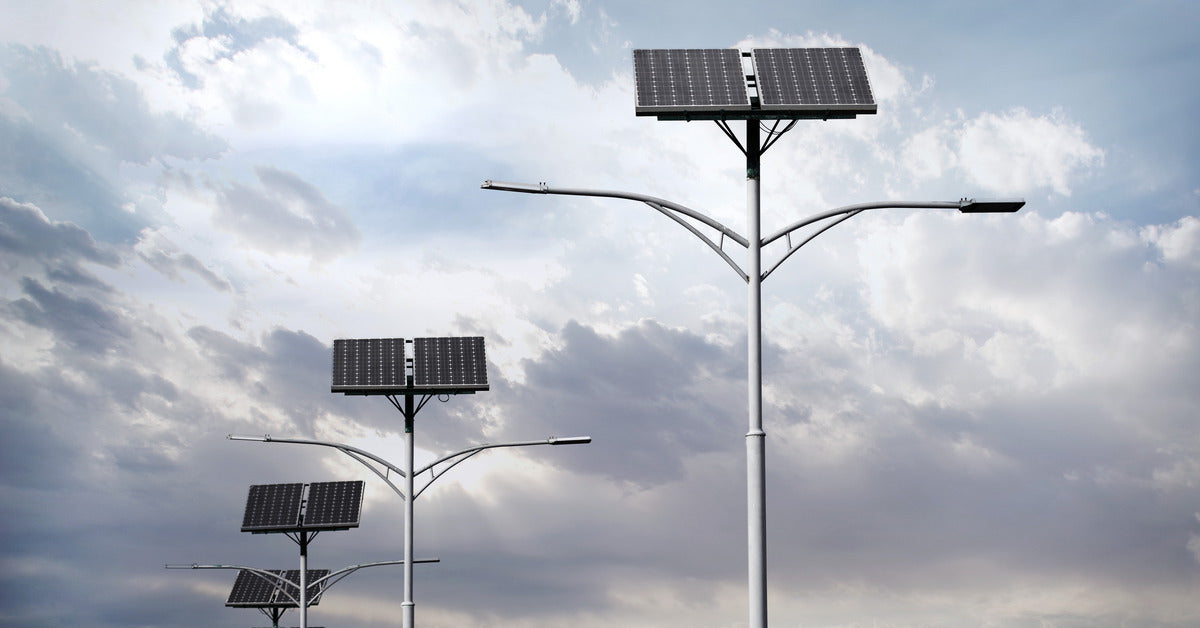 Six tall streetlights with two angled solar panels on the top of the pole. The sky is gray behind the lights.