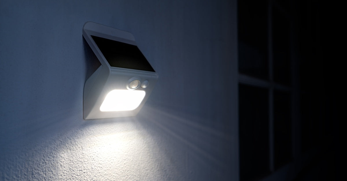 A small solar-powered light mounted to an outside wall of a house next to a darkened window. The light is on.