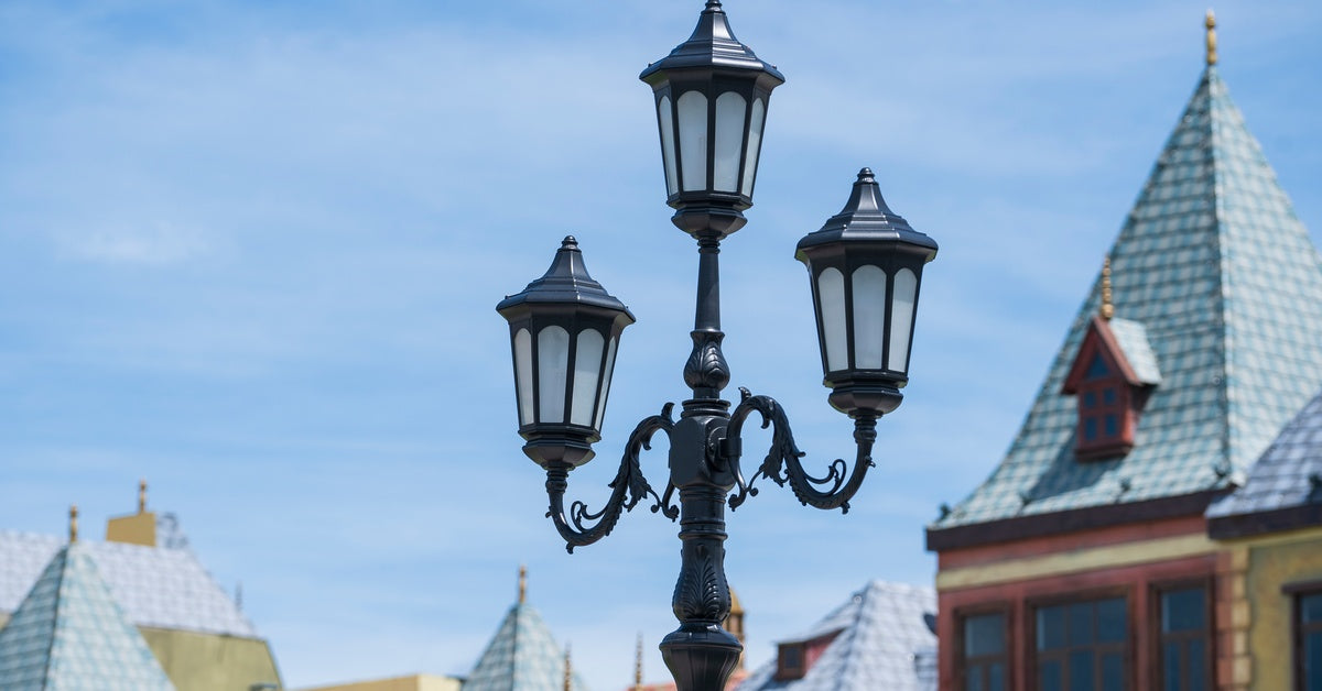 A vintage black lamp post with three lights stands in front of a series of houses in the background.