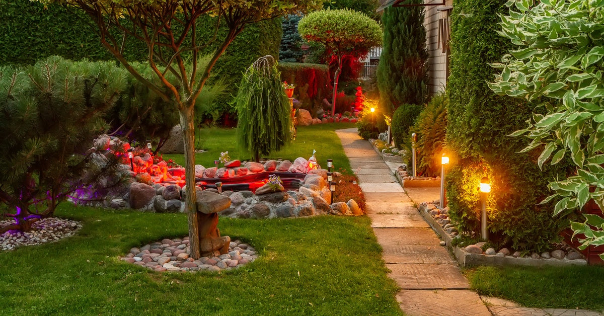 A narrow stone pathway lines the back of a house surrounded by trees and other greenery near a small water feature.