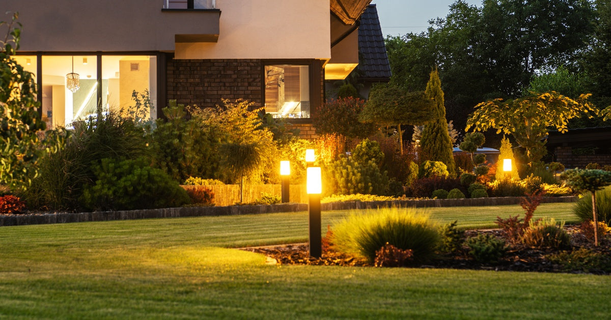 Lights shine bright in the backyard of a home's landscape at night. The lawn looks recently trimmed.