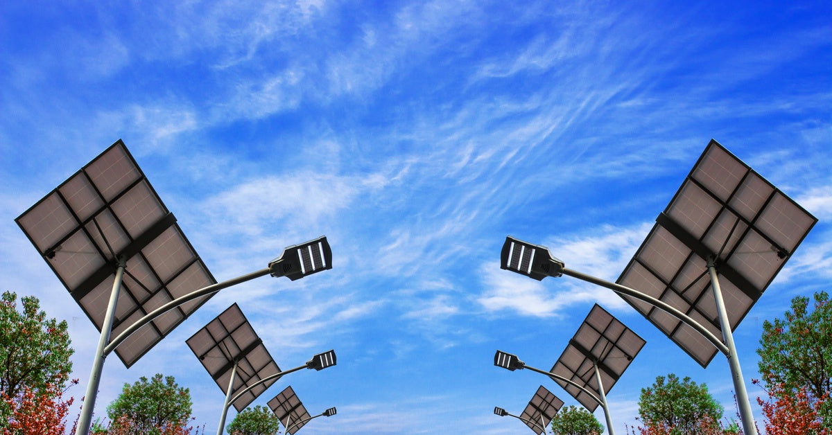 Solar street lamps stretch high up into the sky, with their panels behind them. Several trees stand in the background.
