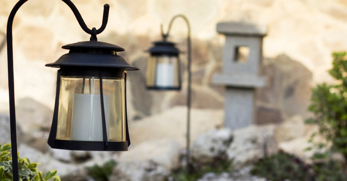 Solar lamps hang from metal hooks in an outdoor garden during the daytime. A miniature stone building sits nearby.