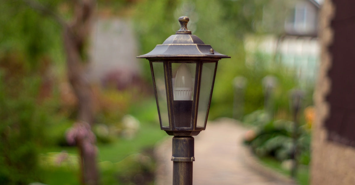A lamp sits on top of a metal post in a backyard. A pathway continues on behind it, and a house sits in the background.
