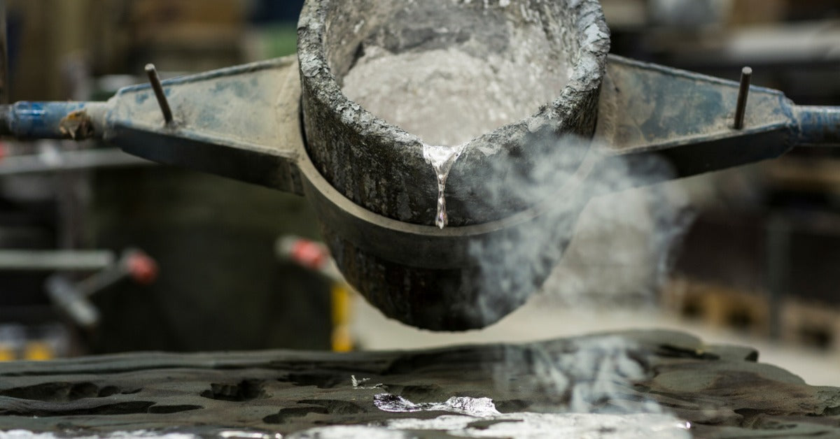 A metal casting ladle pours silvery molten aluminum into a mold as smoke rises across a workshop surface.