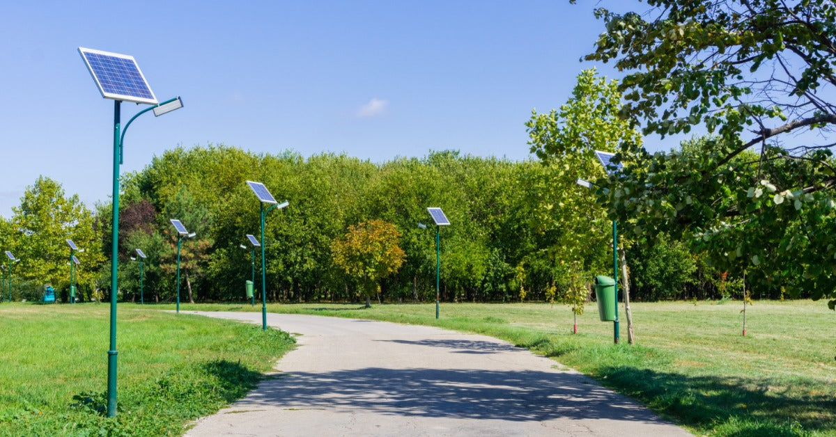 A park features a paved path, solar streetlights, green grass, leafy trees, and a clear blue sky overhead.