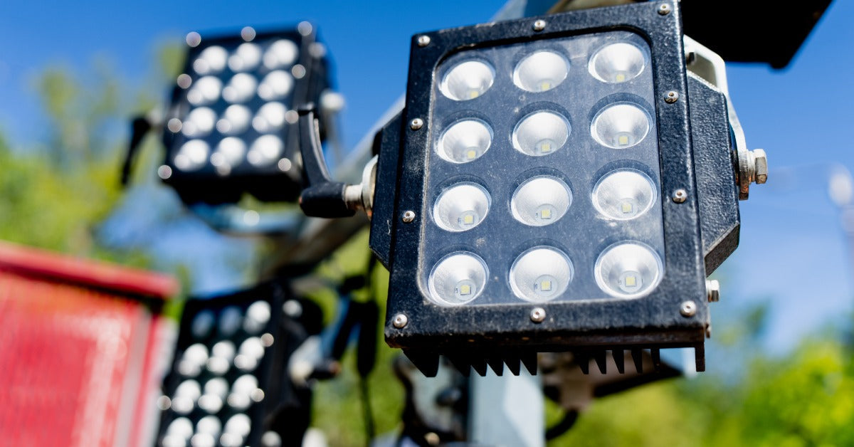 A close-up of black LED floodlight panels mounted on a metal tower, with greenery and blue sky behind them.