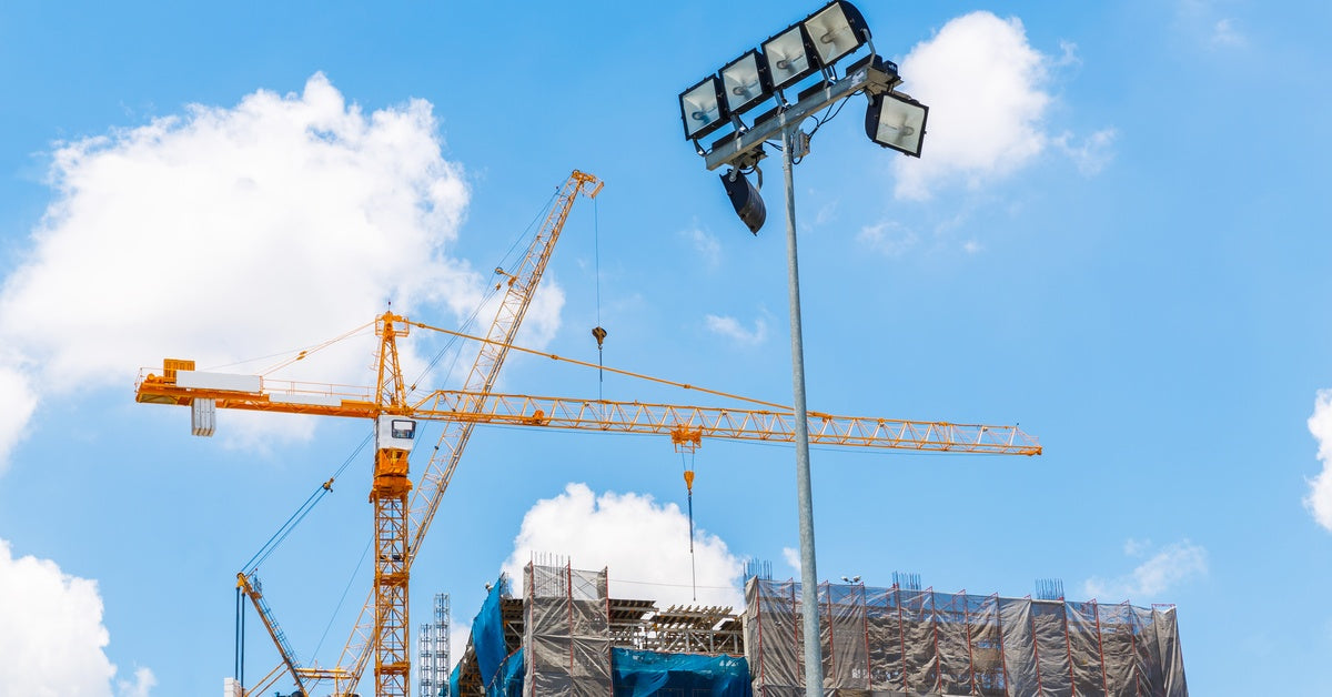 A light tower stands on a construction site. A building is under construction, and a crane stands in the background.