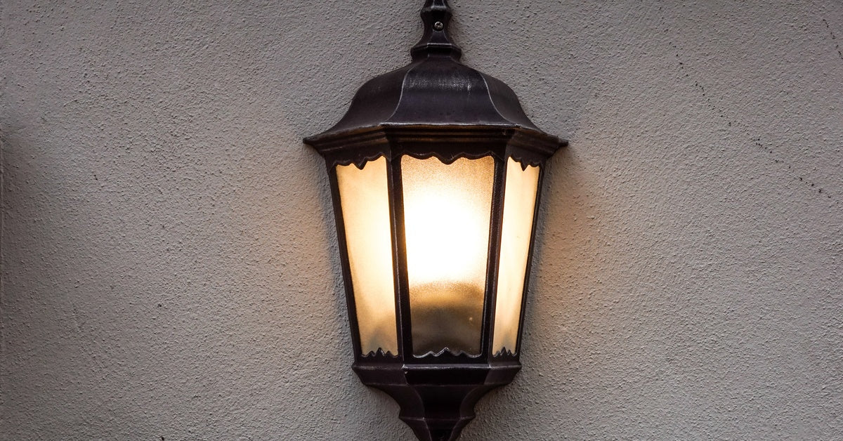 A light is shining inside of a lamp. The lamp is attached to a wall. The wall is white while the lamp is black.