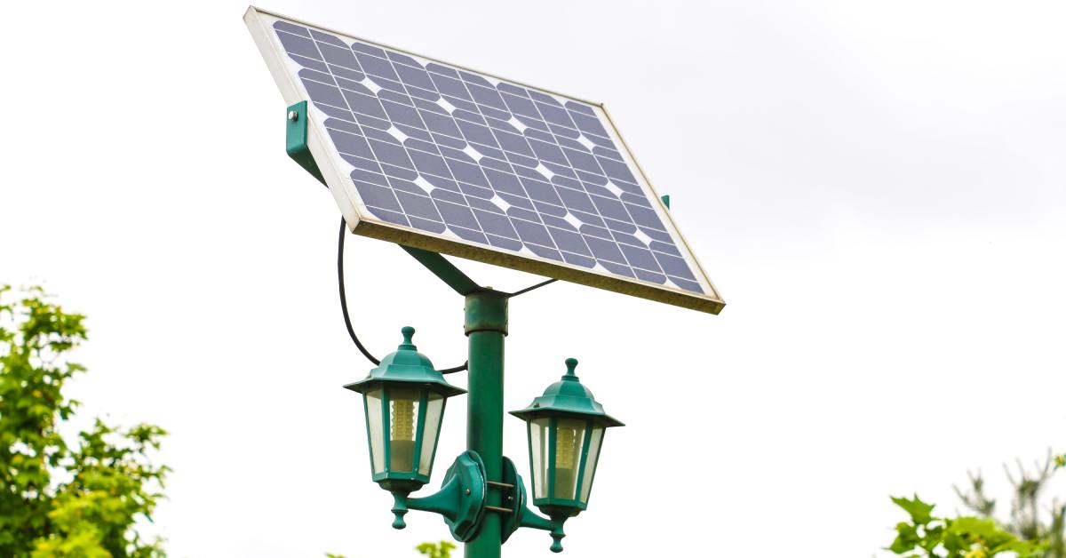 A green solar street light features two lanterns and a large solar panel on top. It stands in an area near trees.