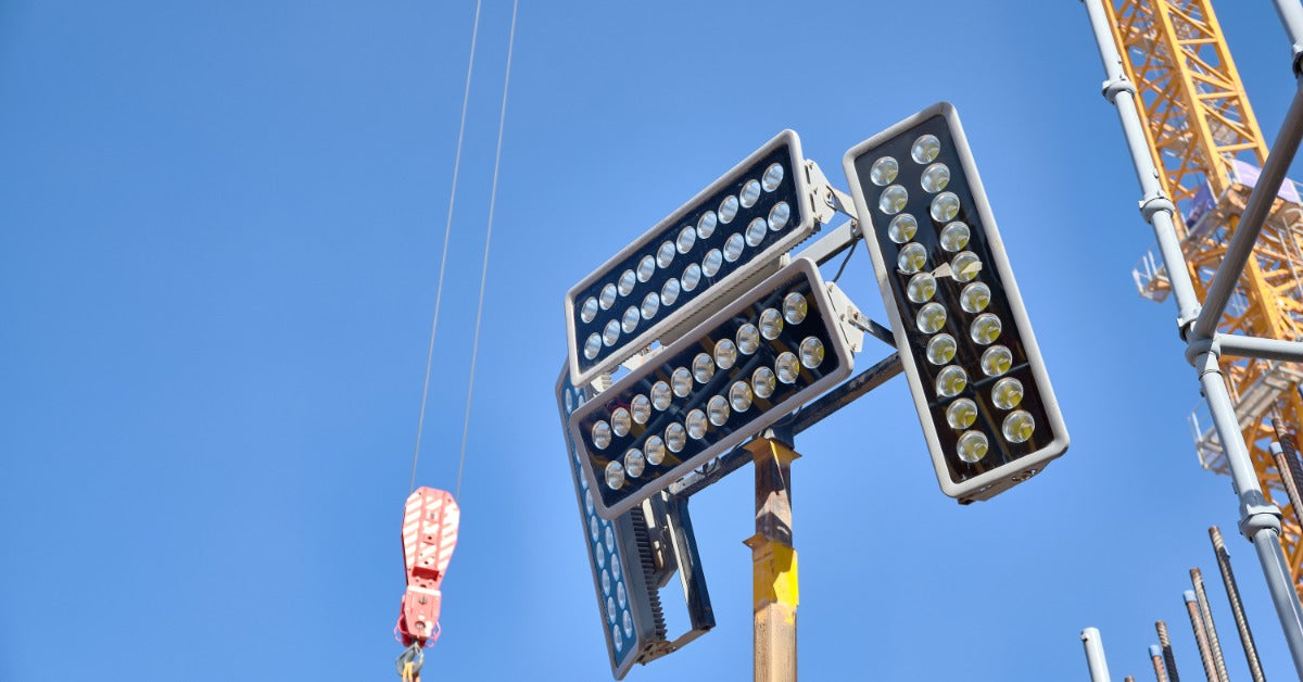 A mobile light tower with multiple bulbs stands tall in the air on a construction site. A crane hook hangs nearby.